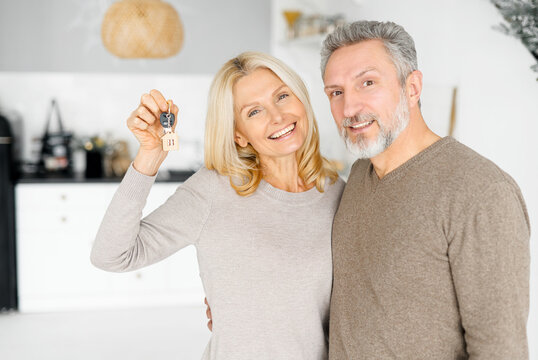 Charming middle-aged wife holding home keys standing in hug of her husband in new apartment, senior couple feel ecstatic about recent purchase of new home