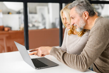 Fototapeta premium Cheerful middle-aged couple spends leisure time online with a laptop at home. Handsome grey-haired man points finger at the screen, beautiful blonde woman watching and laughs. Side view