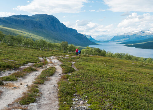 Lapland landscape with beautiful river Lulealven, snow capped mountain, birch tree and footpath of Kungsleden hiking trail with couple of hikers near Saltoluokta, north of Sweden. Summer blue sky