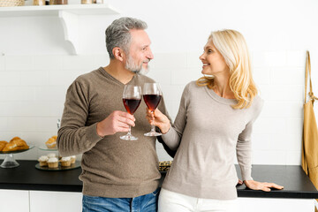 Romantic senior couple spends evening together, standing in the kitchen with glasses of red wine, handsome grey-haired man and charming mid-age woman look to each other, making cheers with glasses