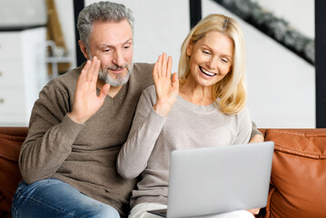 Middle-aged couple is using laptop for video call sitting on comfortable sofa in cozy living room. Cheerful spouses waving into webcam, involved virtual meeting with family. Online communication