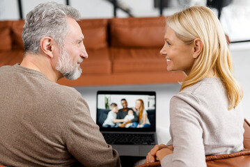 Middle-aged couple using laptop computer for video connection with a grown daughter and her multiracial family, grey-haired man and woman talking online with mixed-race family on computer screen