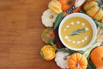 Bowl of butternut squash soup garnished with cream and rosemary, surrounded by various pumpkins, gourds, ornamental corn, and herbs on wood background with copy space.