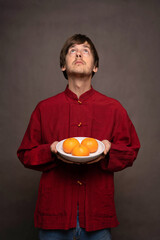 Young handsome tall slim white man with brown hair looking up while holding bowl of oranges in red shirt on grey background