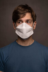 Young handsome tall slim white man with brown hair looking left with face mask on in blue shirt on grey background