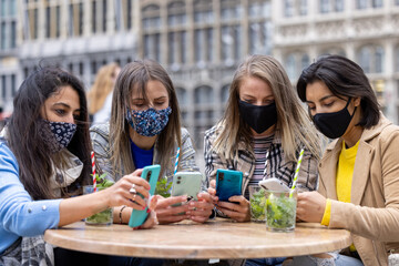 Multiracial young female friends wearing facemasks using mobile smartphone while having a drink outside on the terrace of a cafe in the city during corona virus outbreak - Food and technology concept