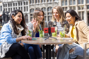 Antwerp, Belgium, May 21, 2021, four mixed race female tourists or students sitting outside in the old city center at a cafe terrace holding a video call using mobile phone. High quality photo