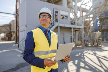 Joyful male engineer in work vest holding notebook and smiling while standing on territory of...