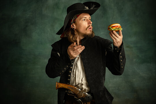 Portrait of one brutal man, medeival pirate with expressive face looking at burger isolated over dark background.