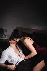 brunette woman in elegant dress kissing man in white shirt on black bedding
