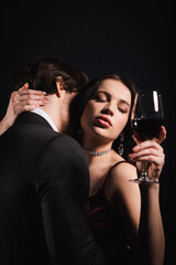 sensual woman with closed eyes holding glass of red wine while man in black suit hugging her isolated on black