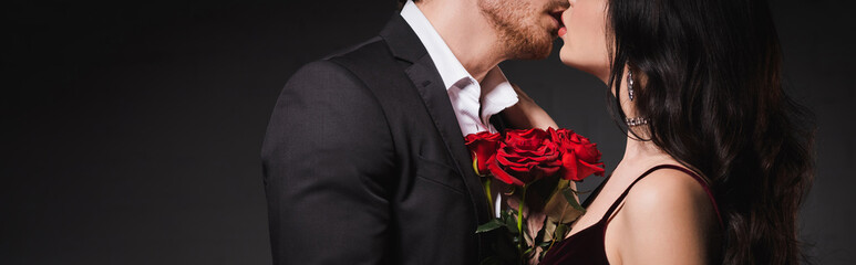 partial view of couple in elegant clothes kissing near red roses on dark background, banner
