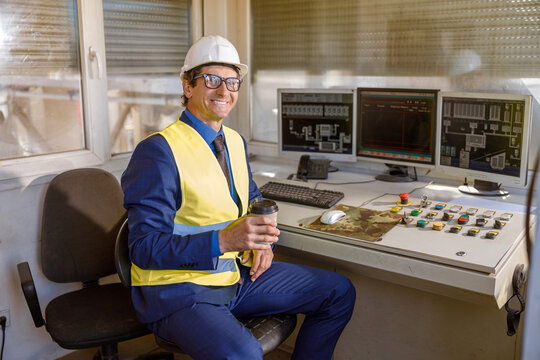 Joyful matured man holding takeaway drink and smiling while sitting at the table with computers and control panel