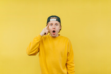 Shocked guy in bright casual clothes and a cap with a shocked face looks at the camera and shows his finger on his temple.