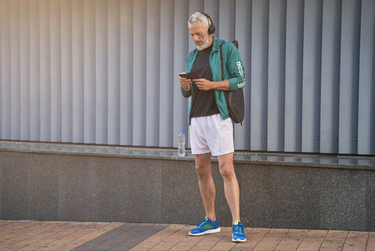 Full length shot of sportive middle aged guy in sportswear and headphones using smartphone, standing outdoors, ready for tennis workout
