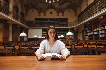 Attractive woman in a white blouse sits at a table in an old atmospheric library and reads a book, looking thoughtfully to the side.