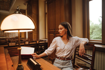 Attractive female student sitting at a desk in a public library and reading a book with a light bulb.