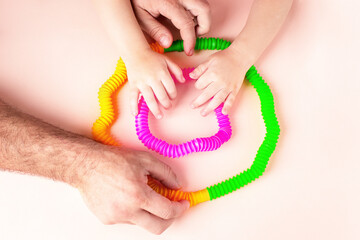 Children's hands are holding a bright antistress toy pop tube toy, next to the father's male hands are helping, pink, green and yellow product. Games with children for motor skills, tactile contact