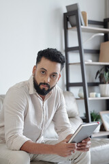 Handsome bearded man with book or planner in hands sitting on sofa and looking at camera