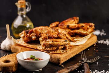 Close-up of BBQ chicken thighs and wings. Appetizing grilled poultry meat, sauce and forks on a wooden cutting board. Dark rustic background.