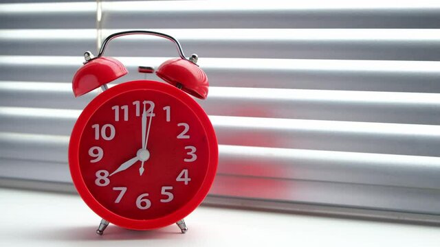 Red alarm clock rang early morning eight clock morning, standing at window against background of closed blinds.