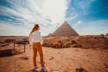Foto op Canvas A girl in orange pants - a tourist stands with her back to the camera and looks at the pyramids. Meditation near the pyramids in Cairo, Egypt © popovatetiana