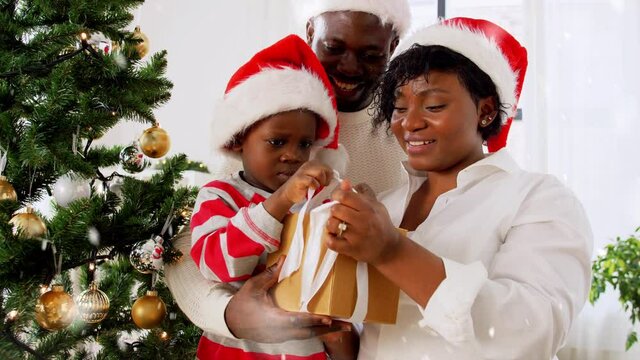 family, winter holidays and people concept - happy african american mother, father and baby son opening gift box at home on christmas over snow