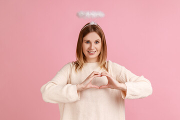Portrait of cute pretty blond angelic woman with nimbus over head looking at camera and showing heart shape with hands, wearing white sweater. Indoor studio shot isolated on pink background.