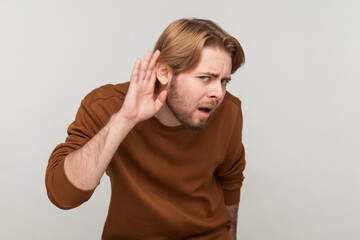 Fototapeta premium Portrait of man with beard wearing sweatshirt, standing with hand on ear and want to hear something, listening to interesting talks and private secrets. Indoor studio shot isolated on gray background.
