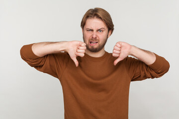 Portrait of man with beard wearing sweatshirt, showing thumbs down, expressing disapproval, criticizing and disagreeing with suggestion. Indoor studio shot isolated on gray background. - obrazy, fototapety, plakaty
