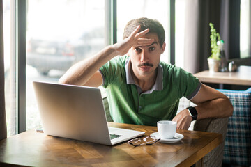 Portrait of man wearing green T-shirt, working online on laptop, holding hand over head, looking...