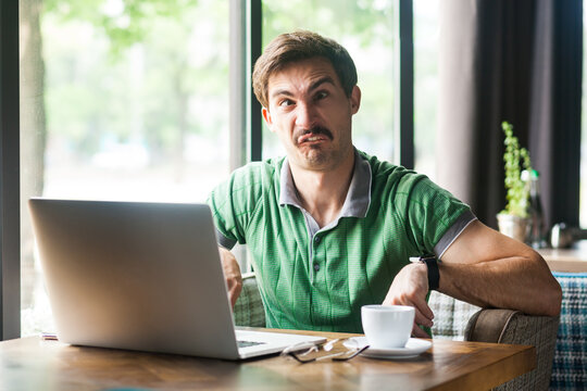Portrait of man employee wearing green T-shirt, crossing eyes, fooling around and having fun, sitting at workplace in front of laptop. Indoor shot near big window, cafe background.