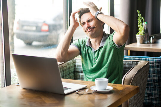Portrait of desperate businessman wearing green T-shirt, feeling sorry and blaming himself for the mistake while working online on laptop, Indoor shot near big window, cafe background.