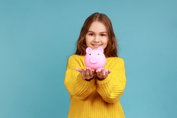 Portrait of smiling cute little girl holding out piggy bank to camera, happy with money savings,...