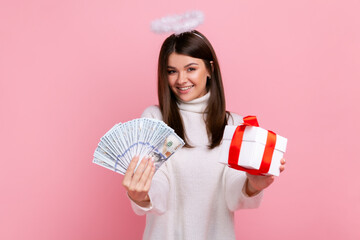 Happy young brunette angelic woman with nimb over head, holding fan of dollar banknotes and gift box, wearing white casual style sweater. Indoor studio shot isolated on pink background.