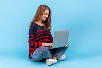 Positive woman wearing striped casual style sweater, sits with crossed legs and working online on lap top, being satisfied with her freelance job. Indoor studio shot isolated on blue background.