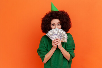 Happy woman with Afro hairstyle wearing green casual style sweater holding covering half of mouth with money, having big sum of money as birthday gift. Indoor studio shot isolated on orange background