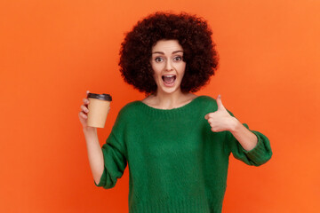 Excited woman with Afro hairstyle wearing green casual style sweater holding coffee to go and showing thumb up, recommend coffee house. Indoor studio shot isolated on orange background.