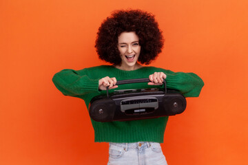 Funny excited woman with Afro hairstyle wearing green casual style sweater holding tape recorder, pushing button, listening to loud music. Indoor studio shot isolated on orange background.