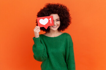 Happy positive young adult woman with Afro hairstyle wearing green casual style sweater covering her eye with social media heart Like icon. Indoor studio shot isolated on orange background.