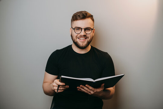 Smiling bearded man hold an notebook in his hands looking at camera. Caucasian man wearing eyeglasses isolated on blue white background. Portrait. Education