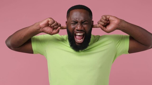 Displeased irritated angry young bearded african american man 20s wears green t-shirt closed eyes cover ears do not want to listen scream isolated on plain pastel light pink background studio portrait