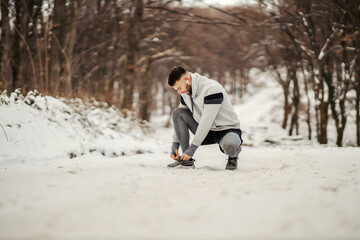 Fit sportsman kneeling on snowy path in nature and tying shoelace. Winter fitness, outdoors sport, sportswear