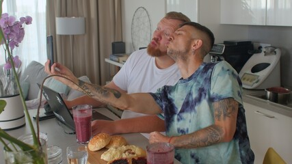 Two gay men drinks a delicious fruit smoothie during breakfast, use laptop and phone in the home kitchen. Vitamin diet to boost immunity. Eating for a healthy weight.