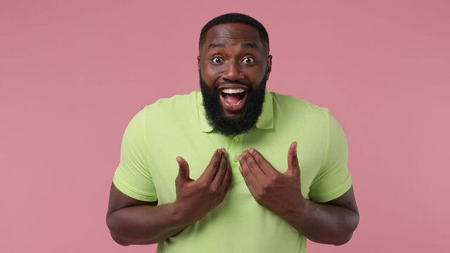 Smiling good kind surprised vivid young bearded african american man 20s wears green t-shirt ask who me oh it so sweet put hands on chest isolated on plain pastel light pink background studio portrait