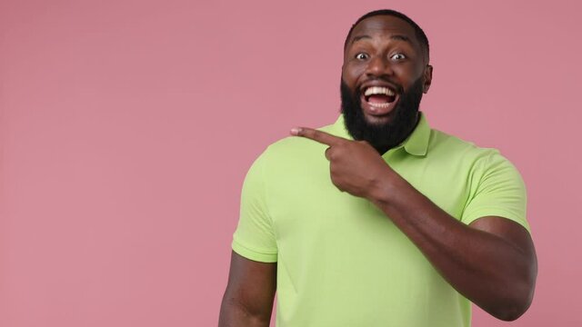 Promoter young bearded american man 20s wears green t-shirt point finger look aside on workspace copy space mockup promo commercial area isolated on plain pastel light pink background studio portrait