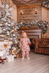 Fototapeta na wymiar a two-year-old girl in a costume with Christmas print stands near the Christmas tree