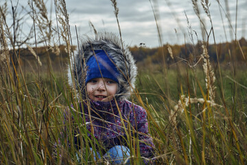 A girl child in a purple jacket and a blue hat sits in the grass and looks straight. Autumn, dry grass and leaves, thick clouds in the sky