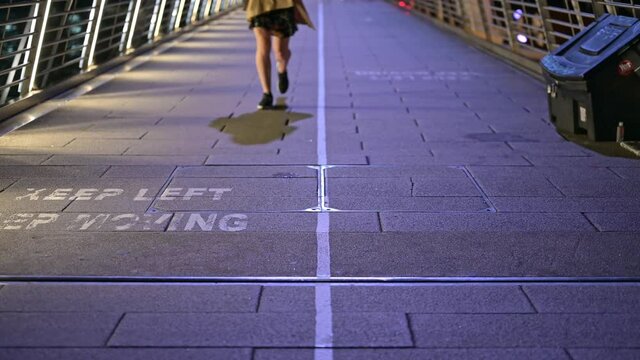 A woman's legs walking the wrong way across a KEEP LEFT sign painted on Jubilee Foot Bridge at night