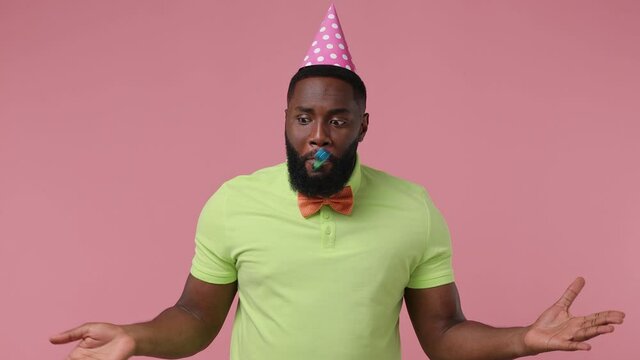 Joyful excited blithesome charismatic stunning young bearded african american man 20s wears green t-shirt bow tie birthday hat blow pipe isolated on plain pastel light pink background studio portrait